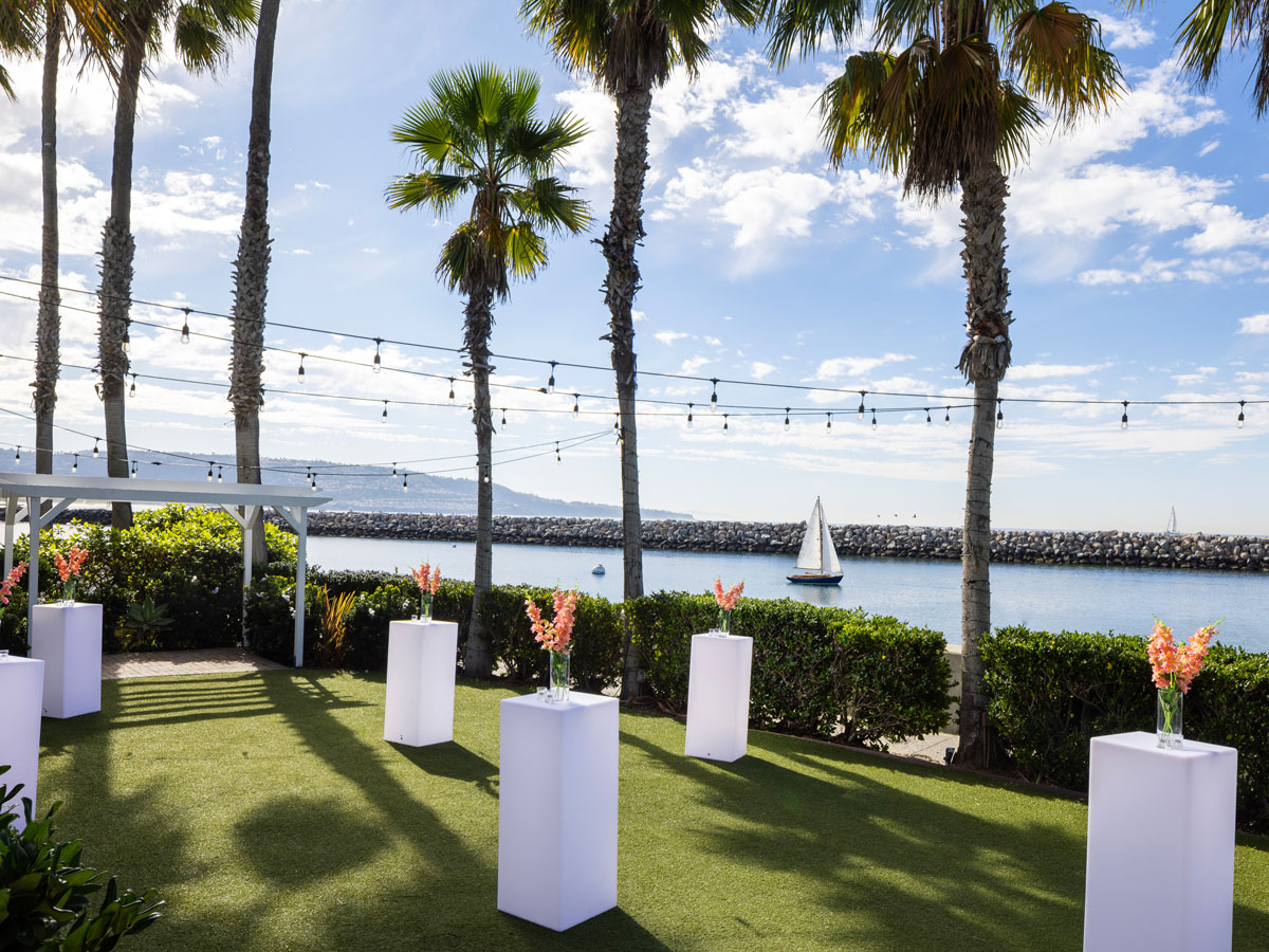 Outdoor event space with white pedestal tables topped with floral arrangements, surrounded by palm trees, overlooking water with a sailboat and string lights overhead.
