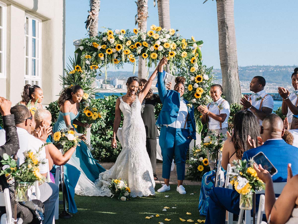 Two brides celebrate under a sunflower arch as guests applaud at an outdoor wedding ceremony by the water.