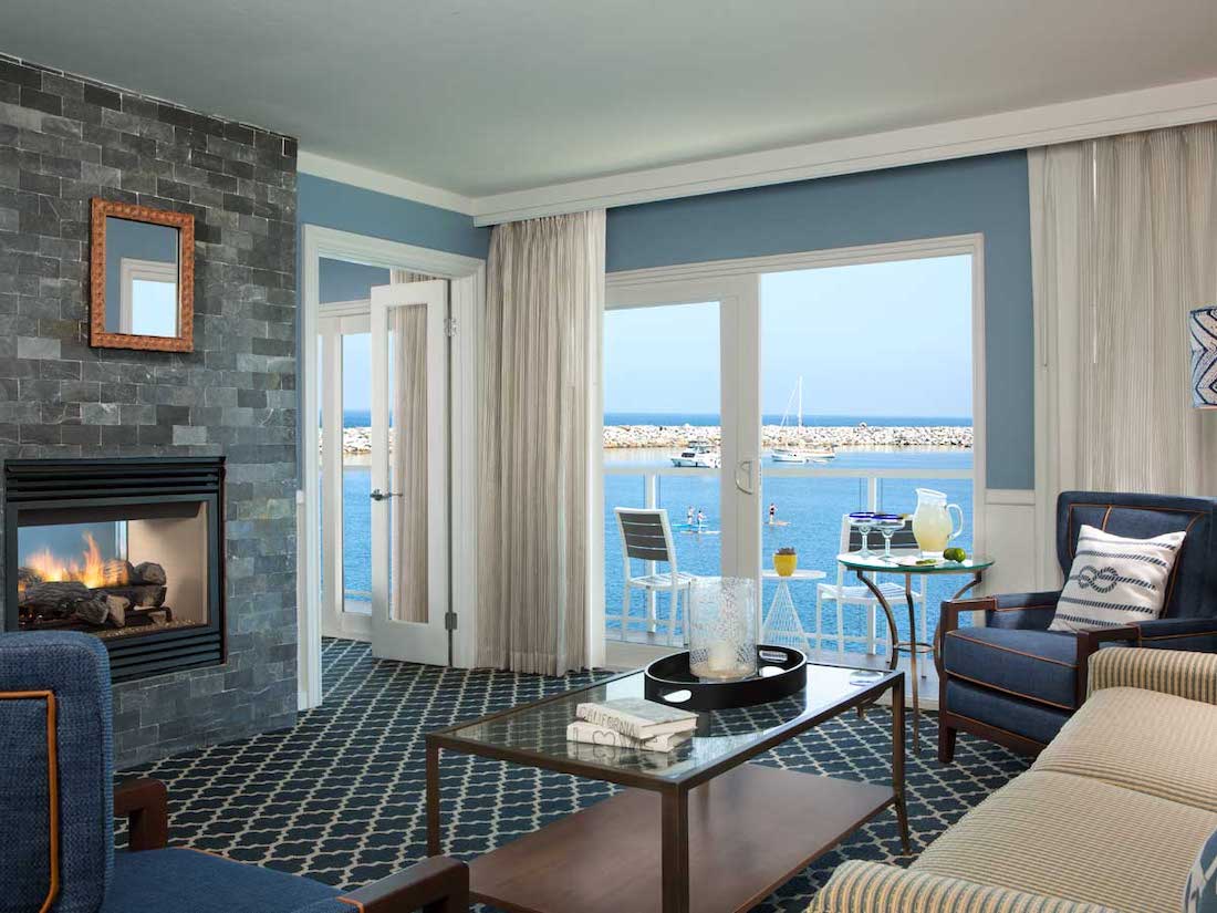 Hotel room with a fireplace, lounge seating, and a glass door leading to a balcony with a view of a marina and boats on the water.