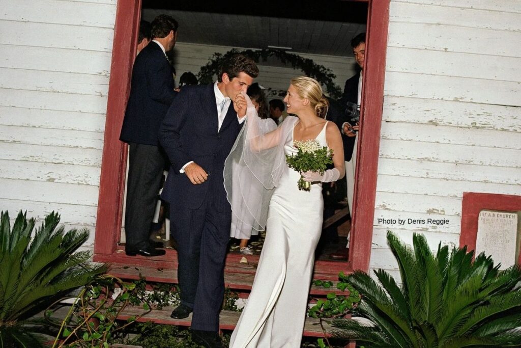 A groom kisses the hand of his bride as they exit a building; she wears a white dress and holds a bouquet, while guests stand in the background.