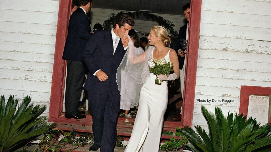 A groom kisses the hand of his bride as they exit a building; she wears a white dress and holds a bouquet, while guests stand in the background.