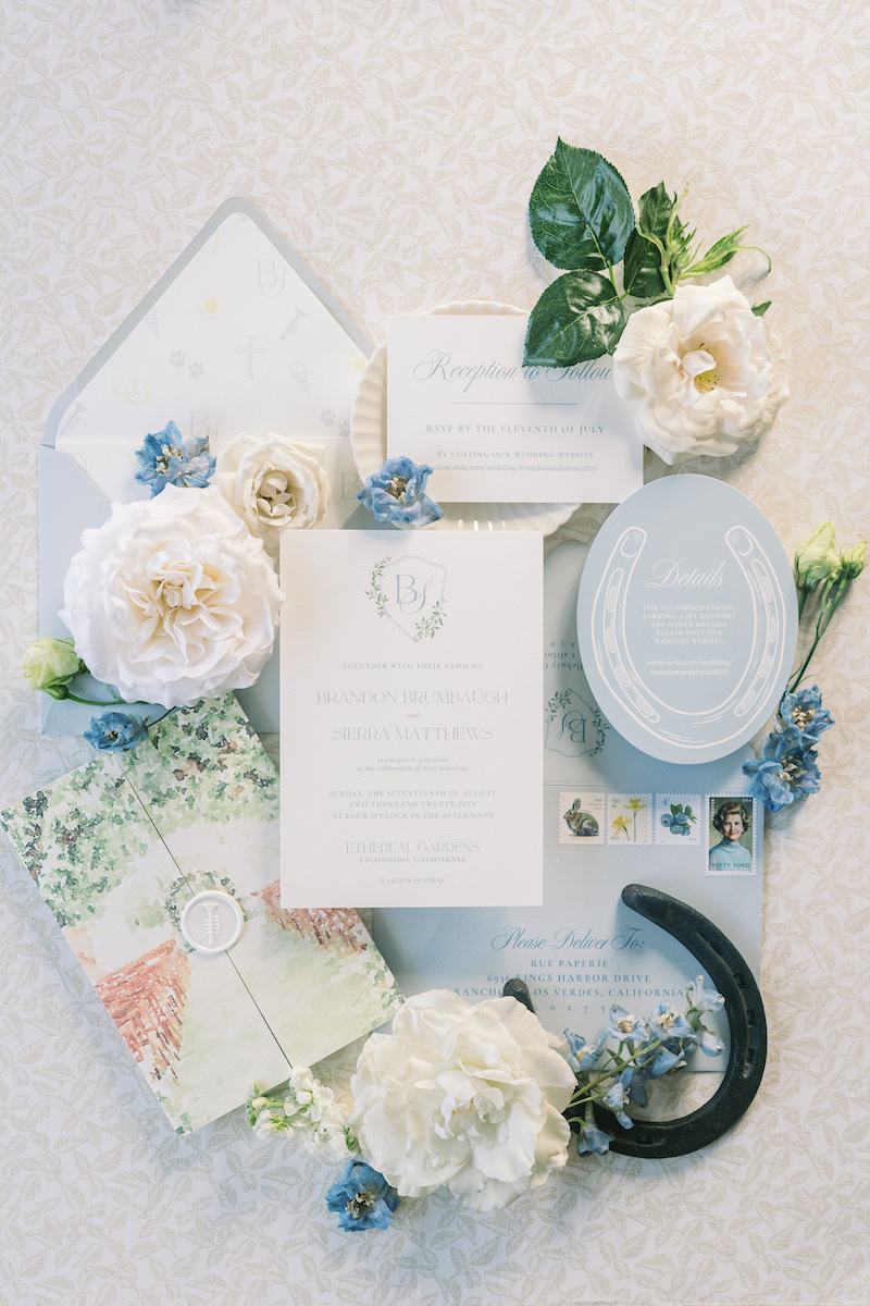 Flat lay of wedding stationery surrounded by white flowers, greenery, blue flowers, and a black horseshoe on a light patterned surface.