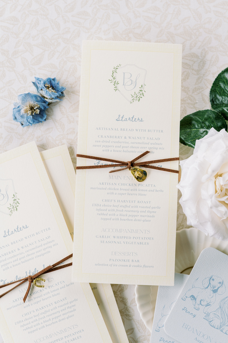 A cream-colored wedding invitation with a brown string and gold heart charm, placed on a textured surface with blue and white flowers.