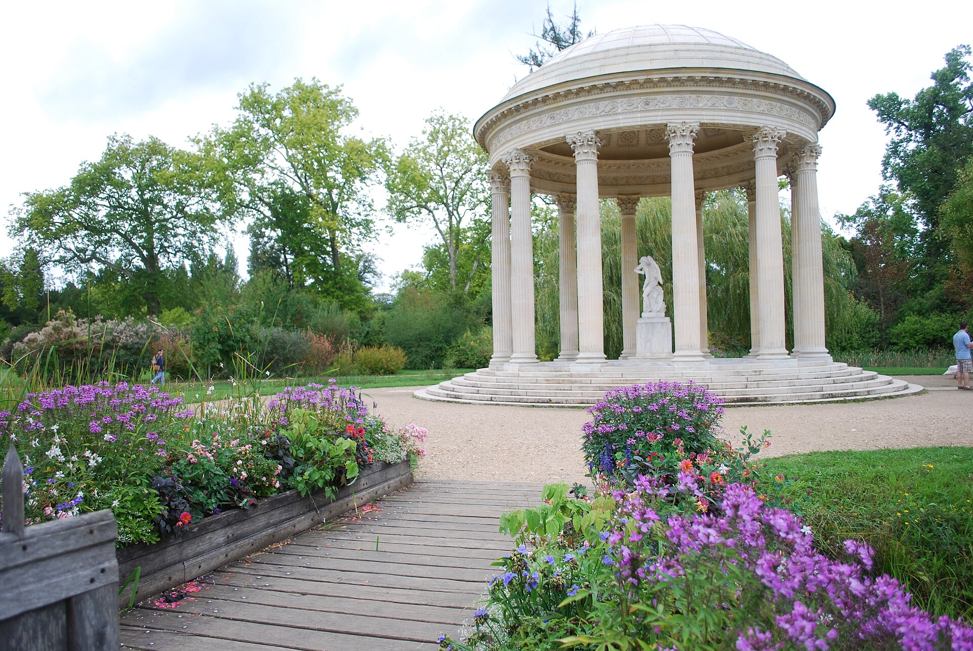 A circular stone pavilion with columns and a statue inside stands in a garden with blooming flowers and greenery.