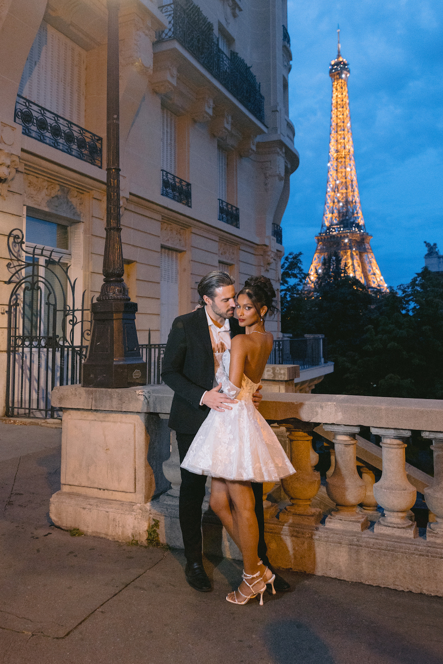 A couple dressed formally poses together near a stone railing with the illuminated Eiffel Tower visible in the background at dusk.