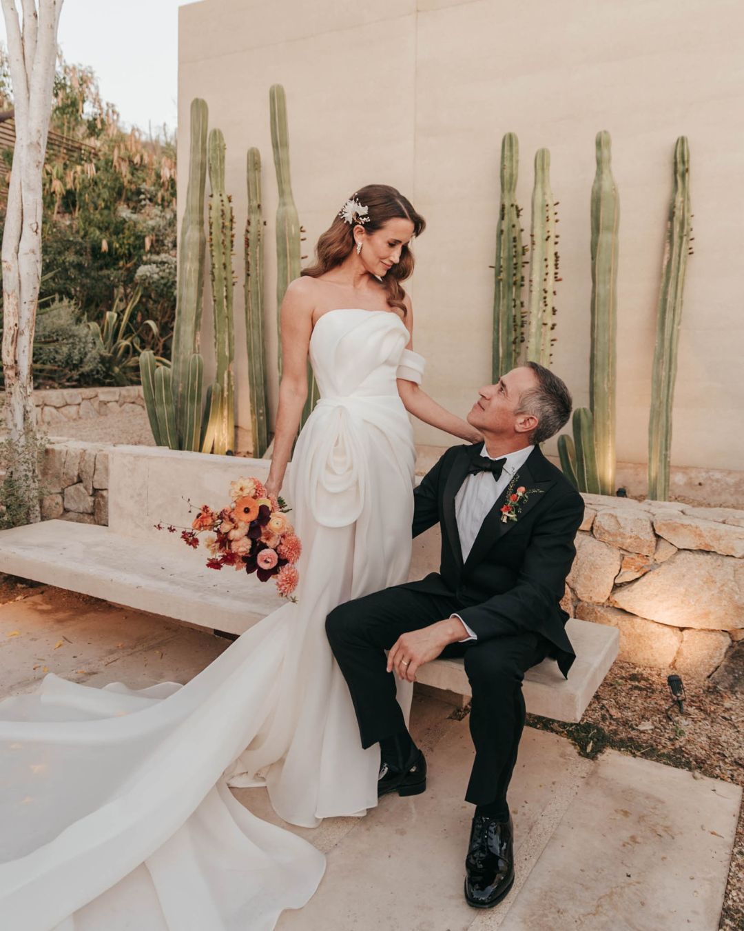 A bride in a white gown stands beside a seated groom in a black tuxedo, with tall cacti and stone walls in the background.