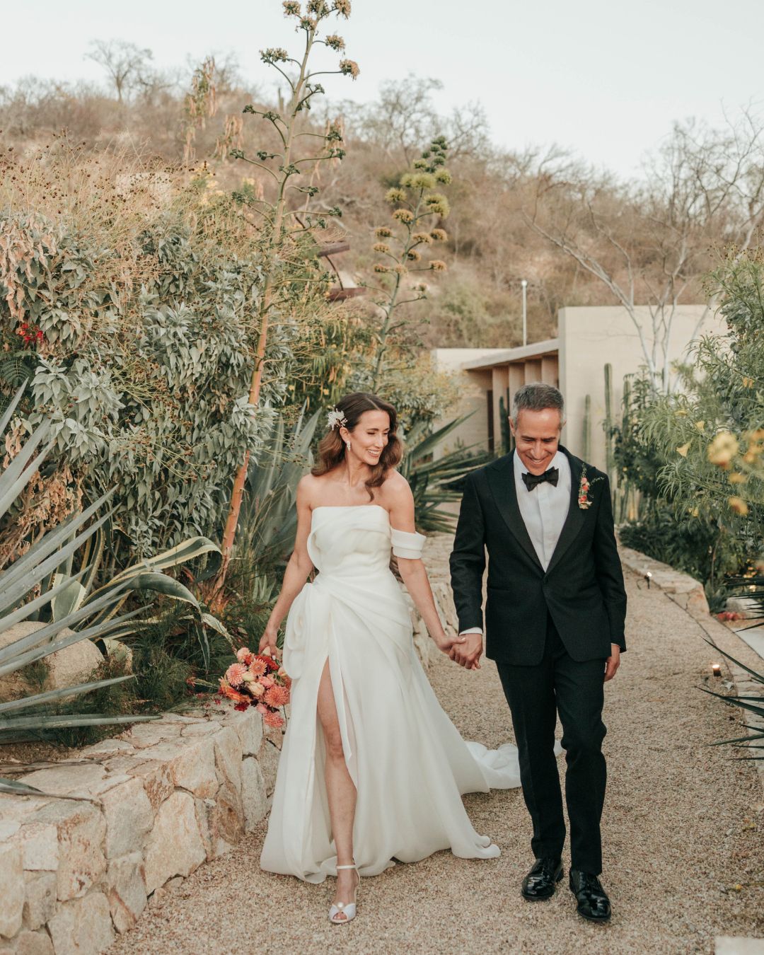 A bride in a white dress and a groom in a black tuxedo walk hand in hand along a gravel path surrounded by plants and stone walls.