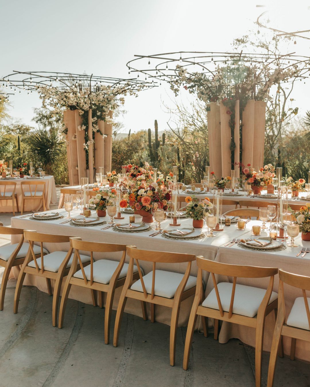 Long banquet tables with wooden chairs are set outdoors, decorated with colorful floral centerpieces and surrounded by tall arrangements and wire frame structures.