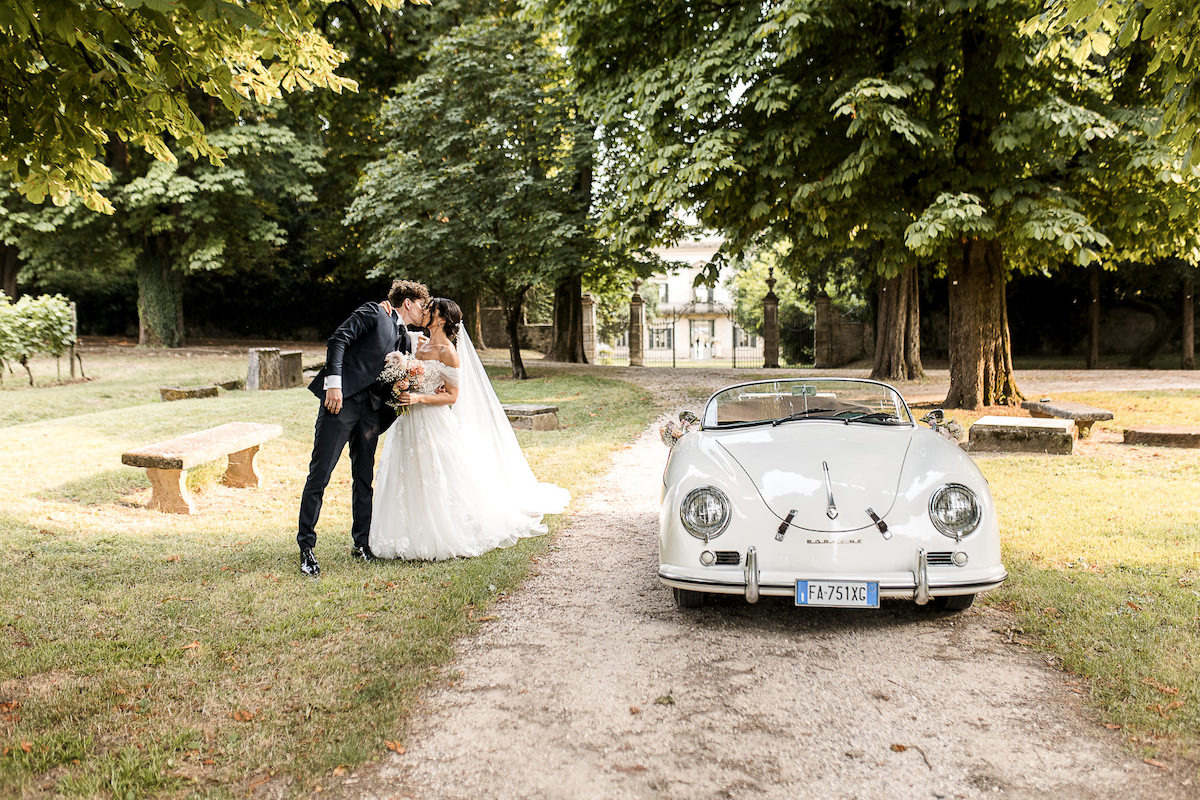 A bride and groom kiss outdoors near a classic white convertible parked on a tree-lined path.