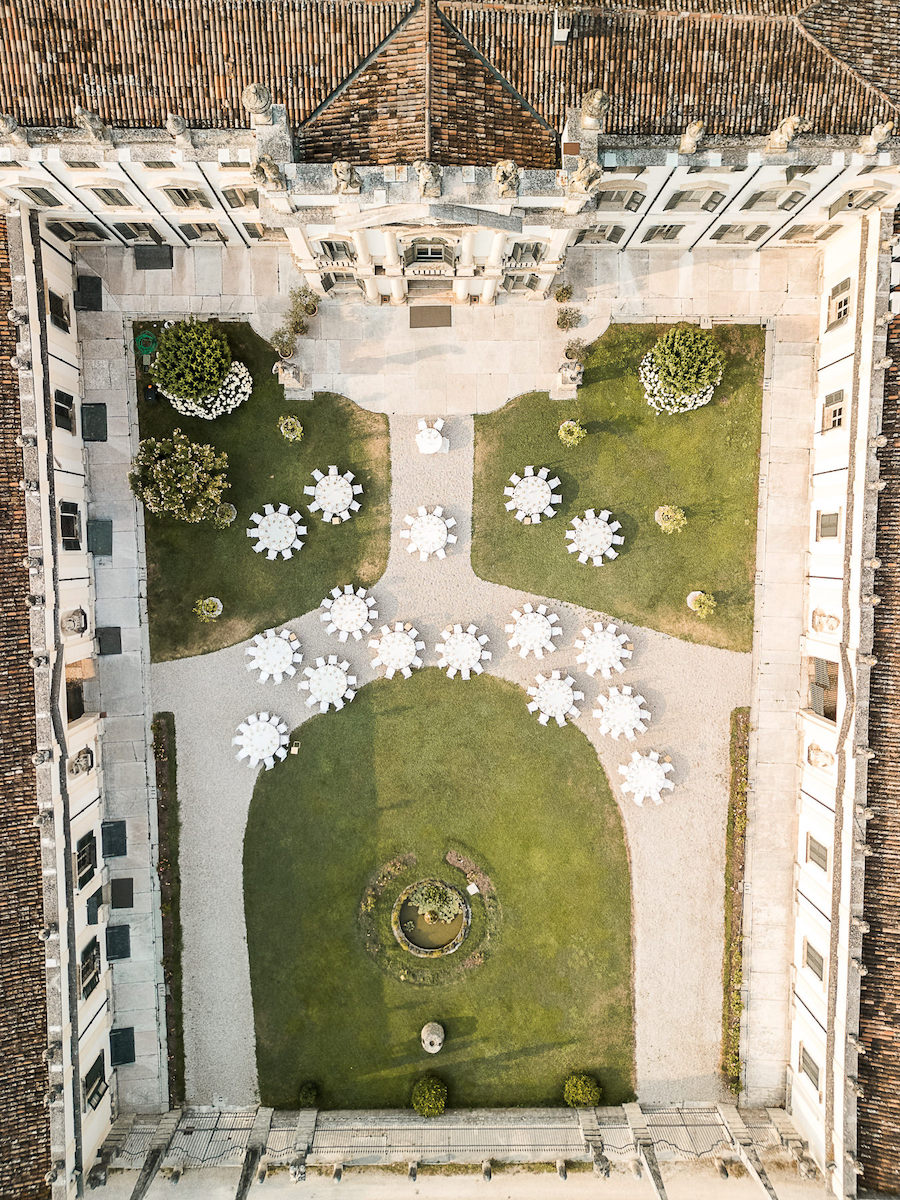 Aerial view of a formal garden with symmetrical pathways, round tables with white umbrellas, and a historic building surrounding the courtyard.