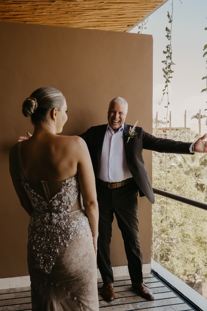 A bride in a formal, embellished dress faces a smiling dad with open arms on a balcony with greenery and city buildings in the background.