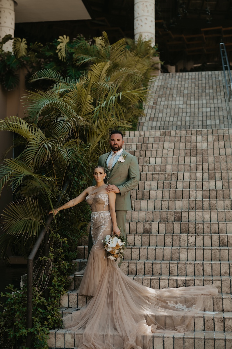 A bride in a beige gown and a groom in a green suit pose on stone steps surrounded by greenery. The bride holds a bouquet and the groom stands behind her.