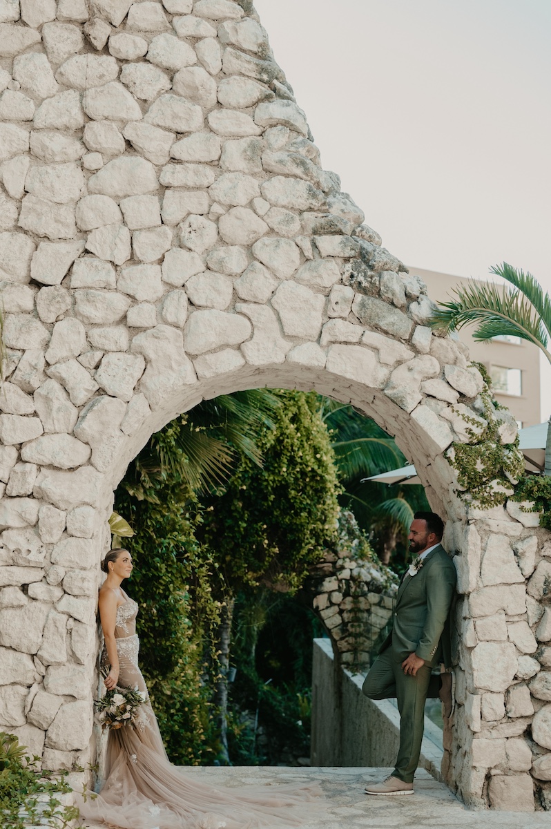 A bride and groom stand facing each other under a stone archway surrounded by greenery.