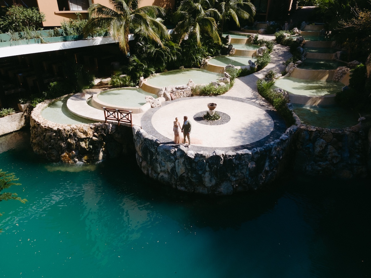Two people stand on a circular stone platform by turquoise water, surrounded by palm trees and terraced pools in a resort setting.