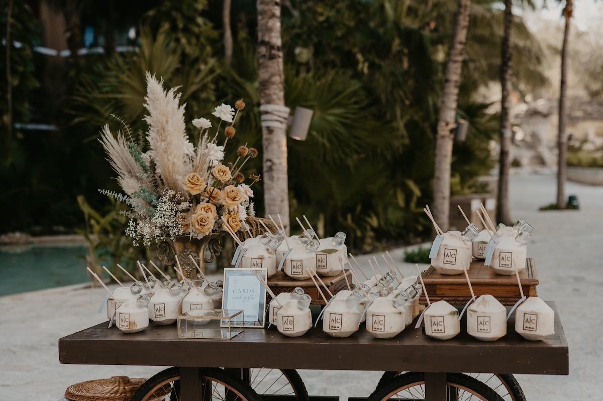 A table displays fresh coconuts with straws and small tags, surrounded by floral arrangements, set outdoors near palm trees.