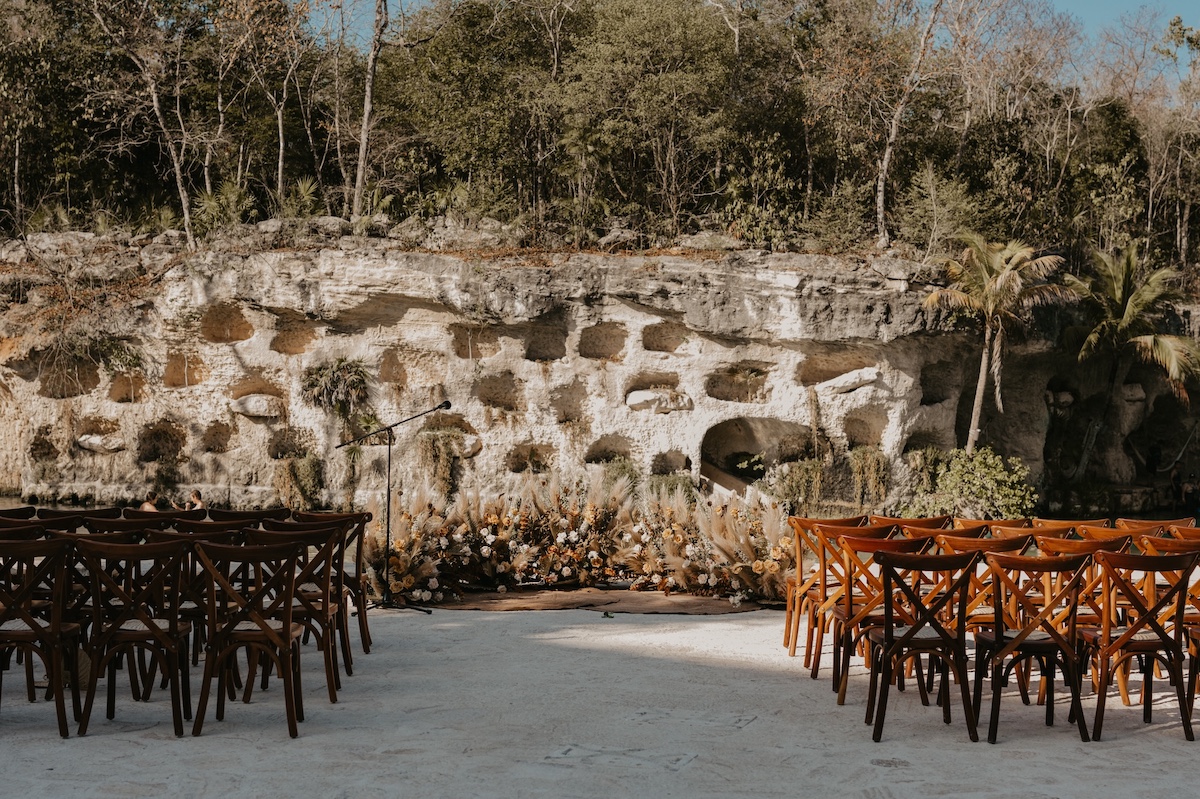 Rows of wooden chairs are arranged outdoors facing a natural rocky wall and greenery, suggesting a ceremony or event setup.
