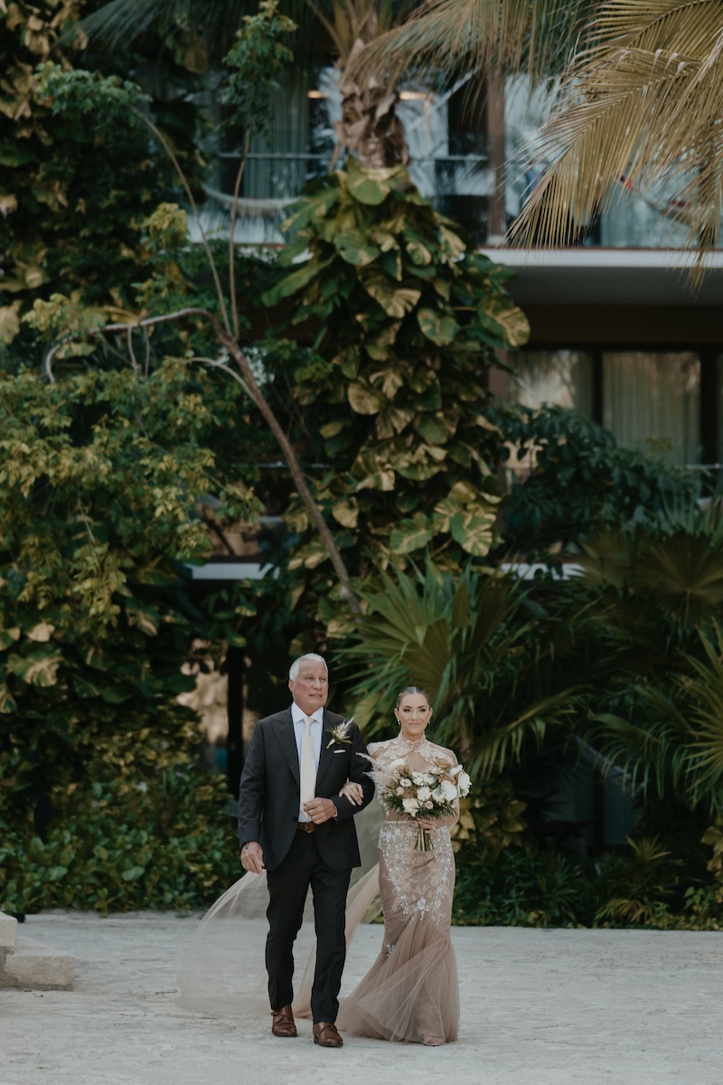 A man in a suit walks beside a woman in a formal gown holding a bouquet, surrounded by lush greenery and modern architecture in the background.