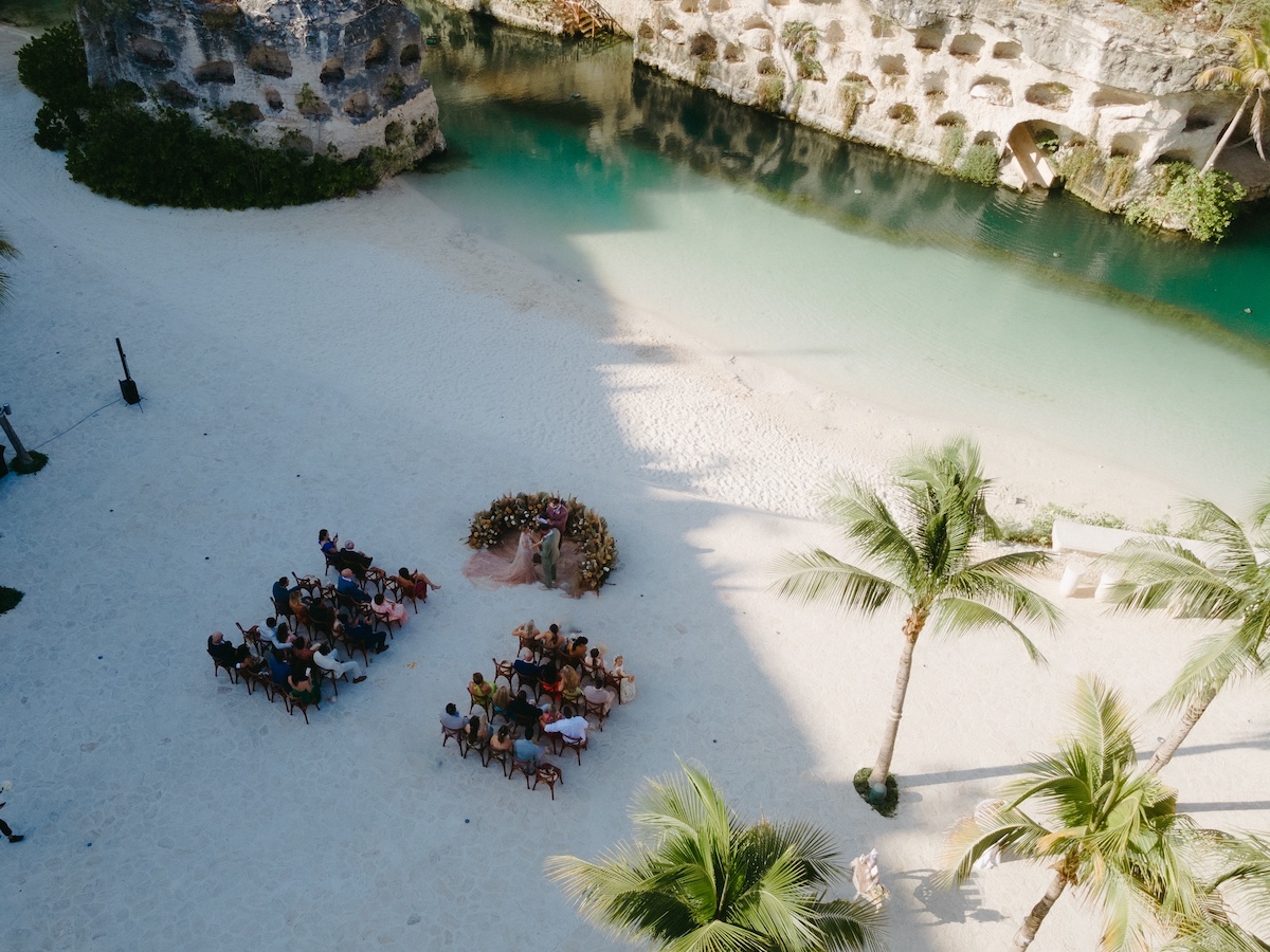 Aerial view of a beachside wedding ceremony with guests seated on sand near palm trees and a turquoise lagoon.