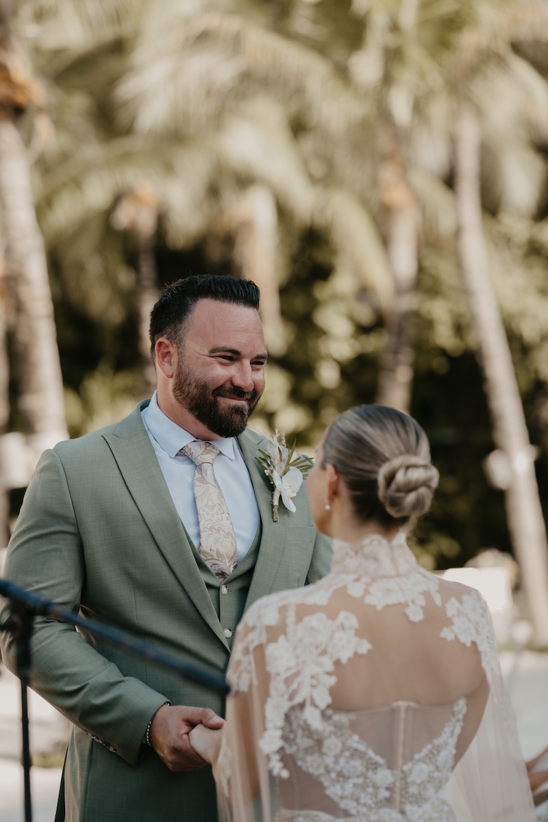 A man in a green suit smiles while holding hands with a woman in a lace wedding dress during an outdoor ceremony with palm trees in the background.