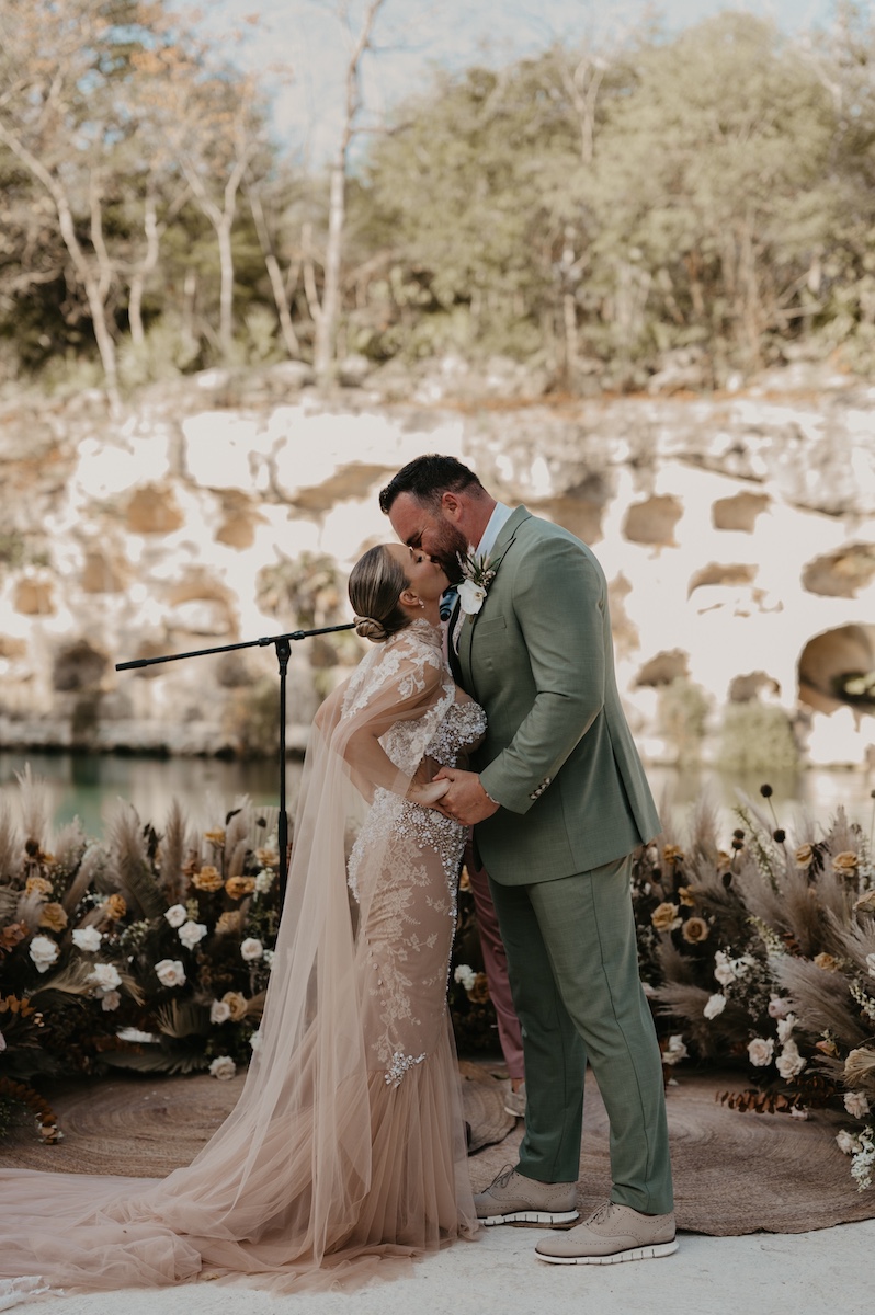 A bride and groom kiss at an outdoor wedding ceremony, surrounded by floral arrangements with a natural rocky background.
