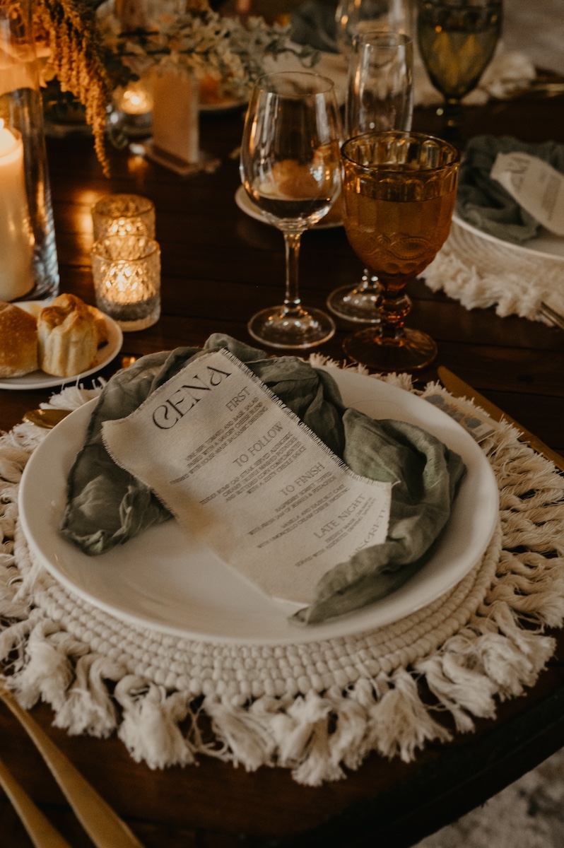 A set dining table with a white plate, printed menu, knotted gray napkin, wine glasses, candles, and bread on a fringed placemat.