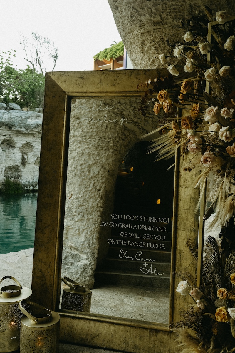 A large mirror leans against a stone wall beside dried flowers and a lantern, reflecting the water and surroundings outside.