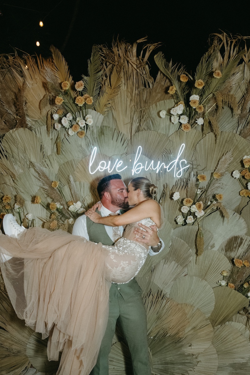 A groom lifts and kisses the bride in front of a dried floral backdrop with a neon sign that reads "love birds.