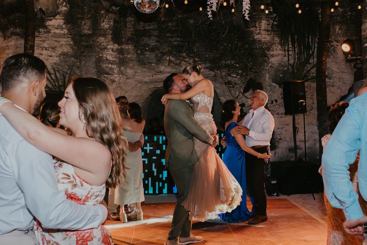 Couples are slow dancing on a warmly lit dance floor; a man lifts a woman in the center while others dance around them in a decorated venue.