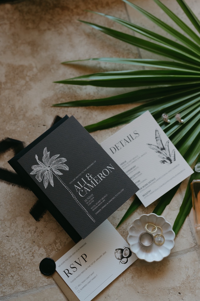 Flat lay of wedding stationery including invitations, RSVP card, and details card, arranged with a palm leaf, two rings in a dish, and a black wax seal on a beige surface.