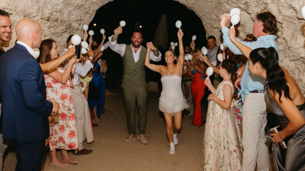 A couple walks hand in hand through a tunnel as guests celebrate, holding white maracas and clapping.