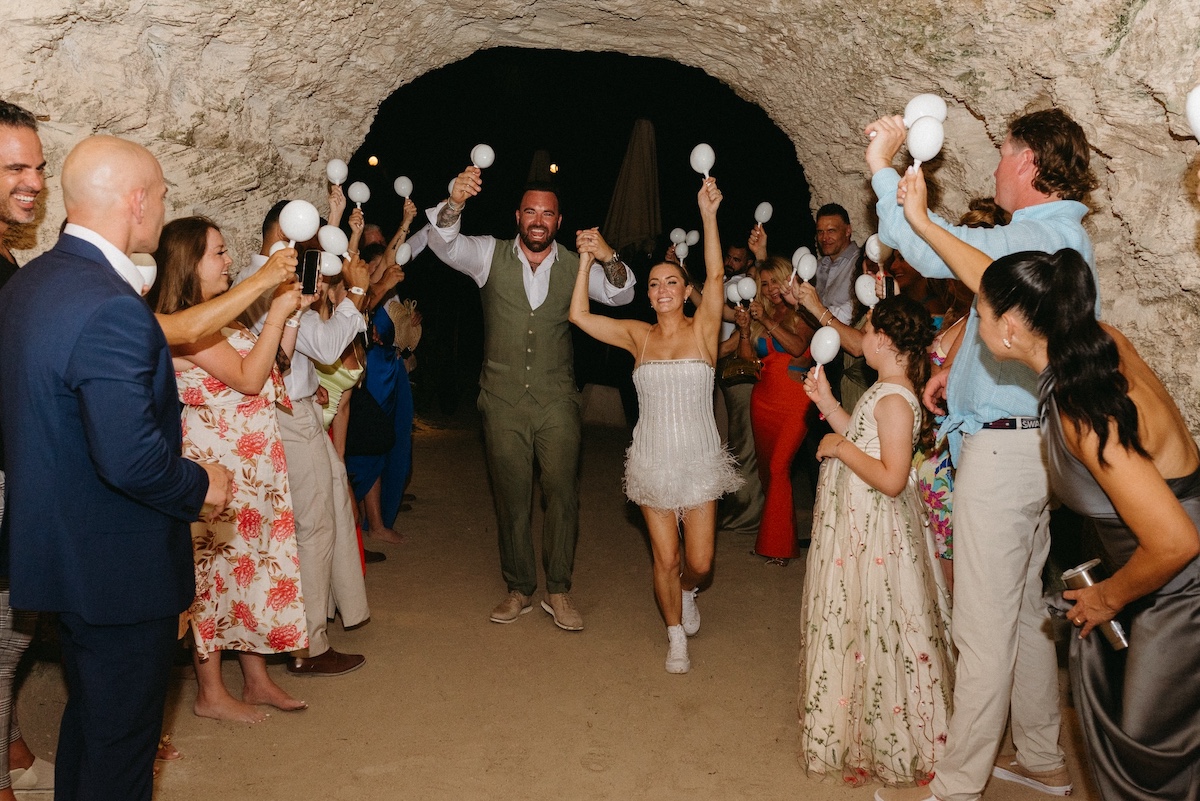 A bride and groom walk through a tunnel while guests on both sides cheer and hold up white decorations.