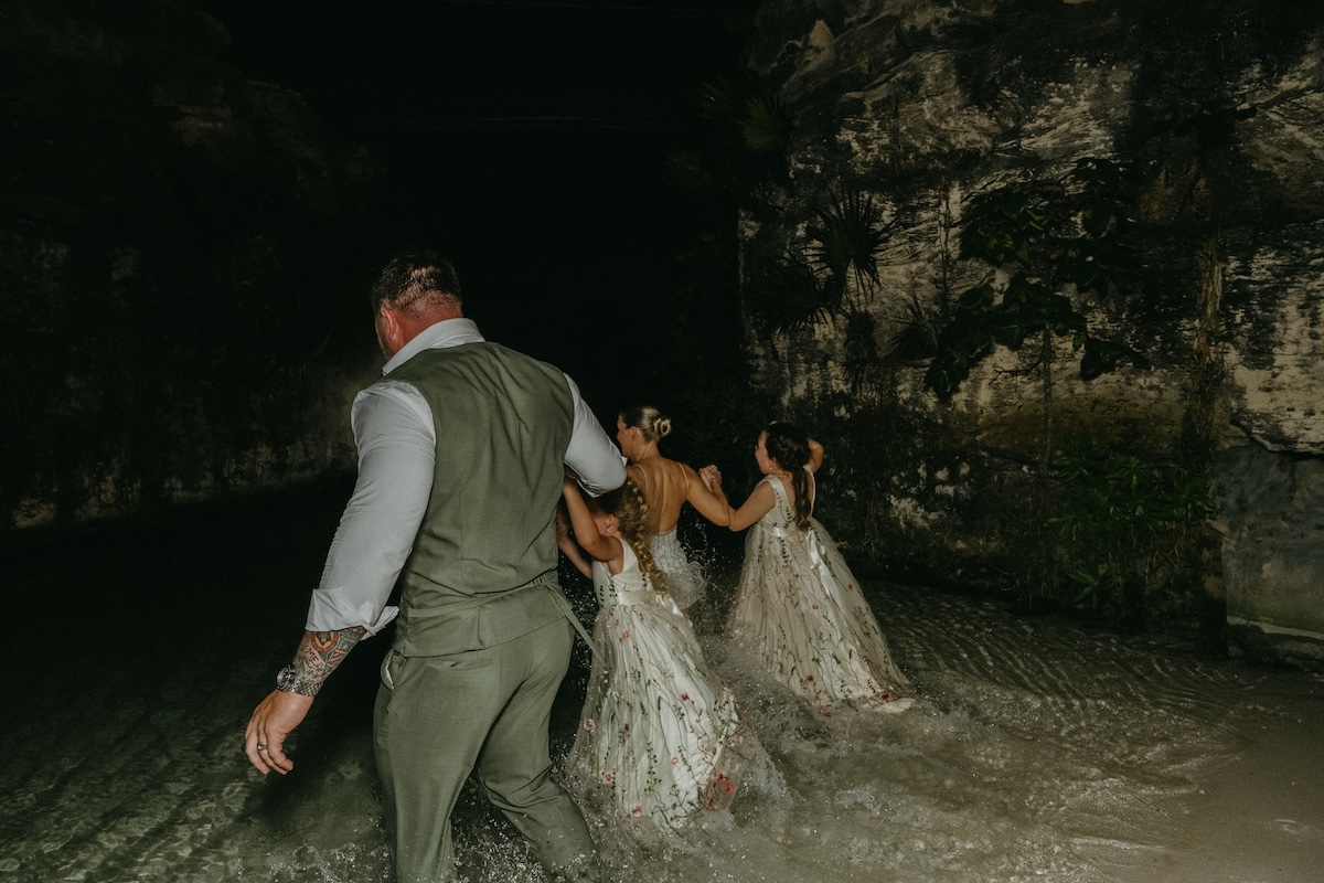 A bride and groom walk with two kids in white dresses walk through shallow water at night near rocky cliffs.