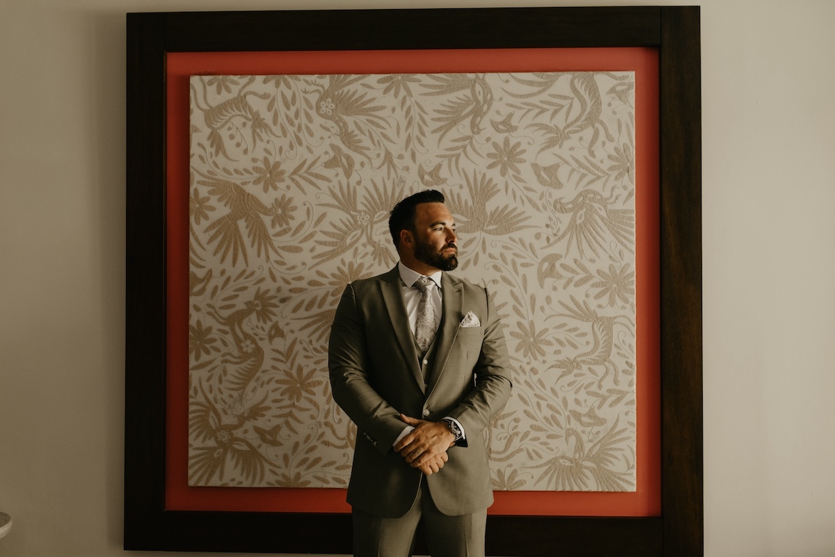 A man in a suit stands in front of a large, patterned artwork framed in red and brown, looking to his left.