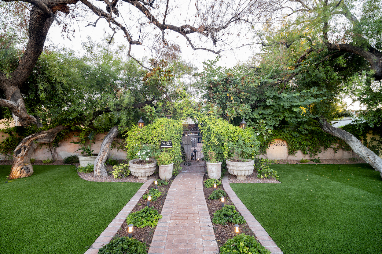A brick walkway leads through a landscaped garden with trimmed grass, large potted plants, and arching trees creating a shaded entryway.
