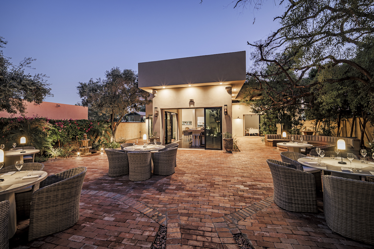 Outdoor dining area with round tables and wicker chairs on a brick patio, surrounded by greenery, trees, and ambient lighting near a modern building at dusk.