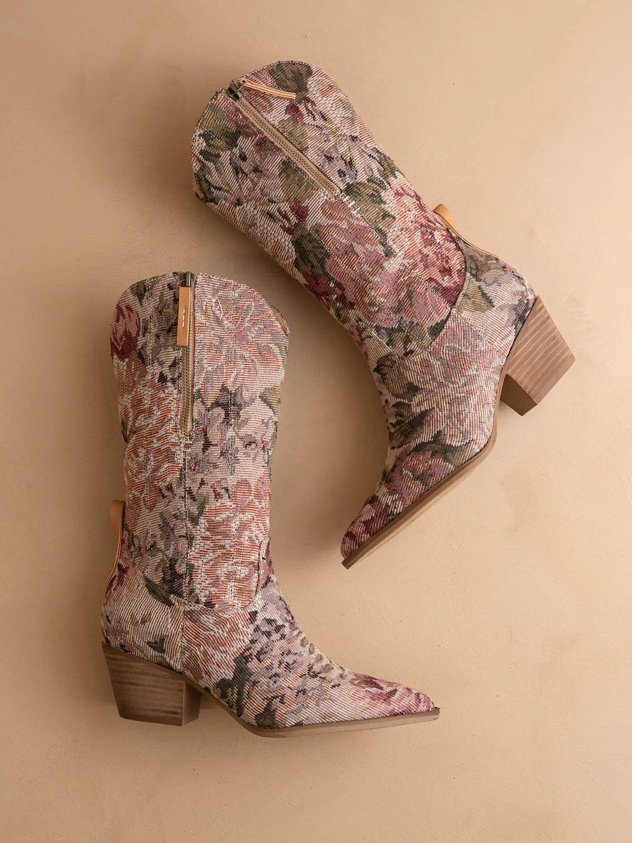 Two floral-patterned cowboy boots with wooden heels are placed side by side on a beige surface, with the inner zipper and pointed toes visible.