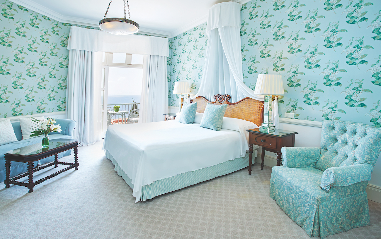 Elegant bedroom with floral wallpaper, large bed with canopy, two nightstands, armchair, sofa, and glass door leading to a balcony with a sea view.