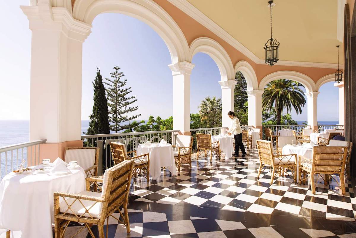 Elegant outdoor restaurant terrace with black-and-white checkered floor, set tables, wicker chairs, large arches, and a view of trees and the ocean; a waiter stands by a table.