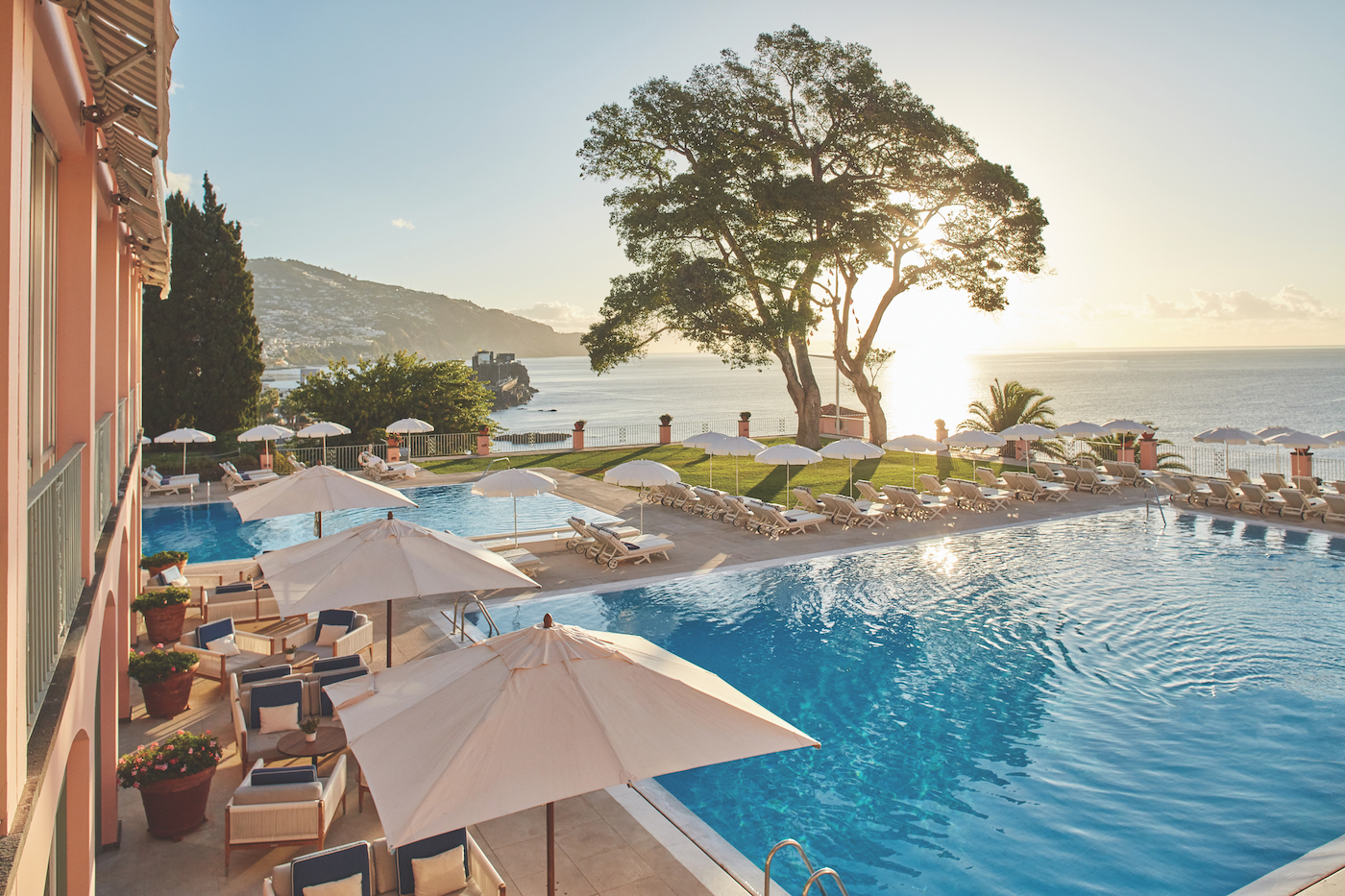 An outdoor pool area with lounge chairs and umbrellas overlooks the ocean and distant hills; the sun is setting behind a large tree.