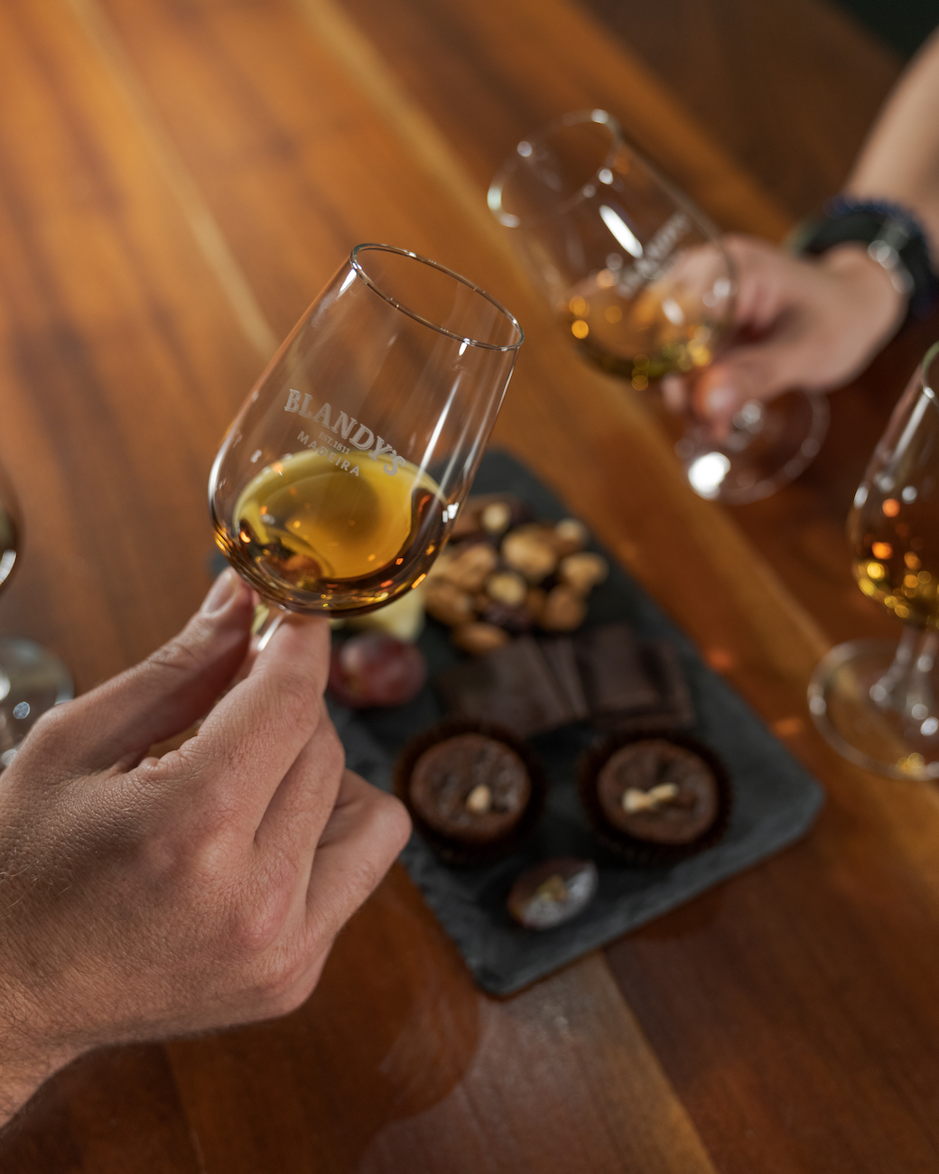Two people hold glasses of amber-colored Madeira, possibly enjoyed during weddings in Portugal, over a wooden table with assorted chocolates and nuts on a slate serving tray.