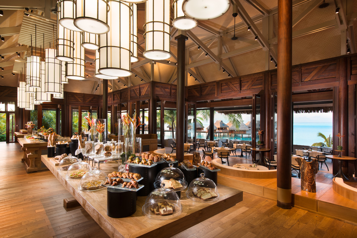 Spacious restaurant with wooden decor, large hanging lights, and a buffet table filled with pastries and desserts; open view to outdoor seating and ocean.