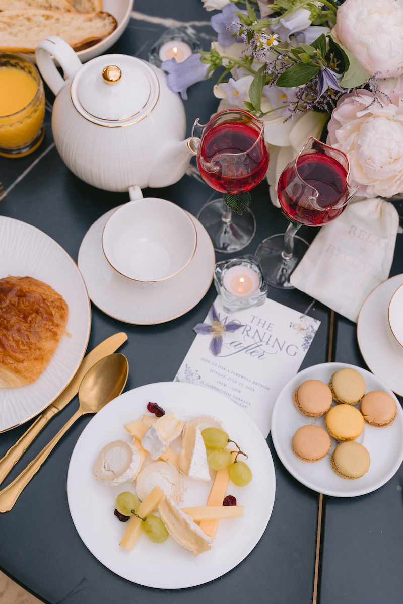 A brunch table with a teapot, two glasses of red wine, a plate of cheese and grapes, macarons, a croissant, orange juice, and flowers.
