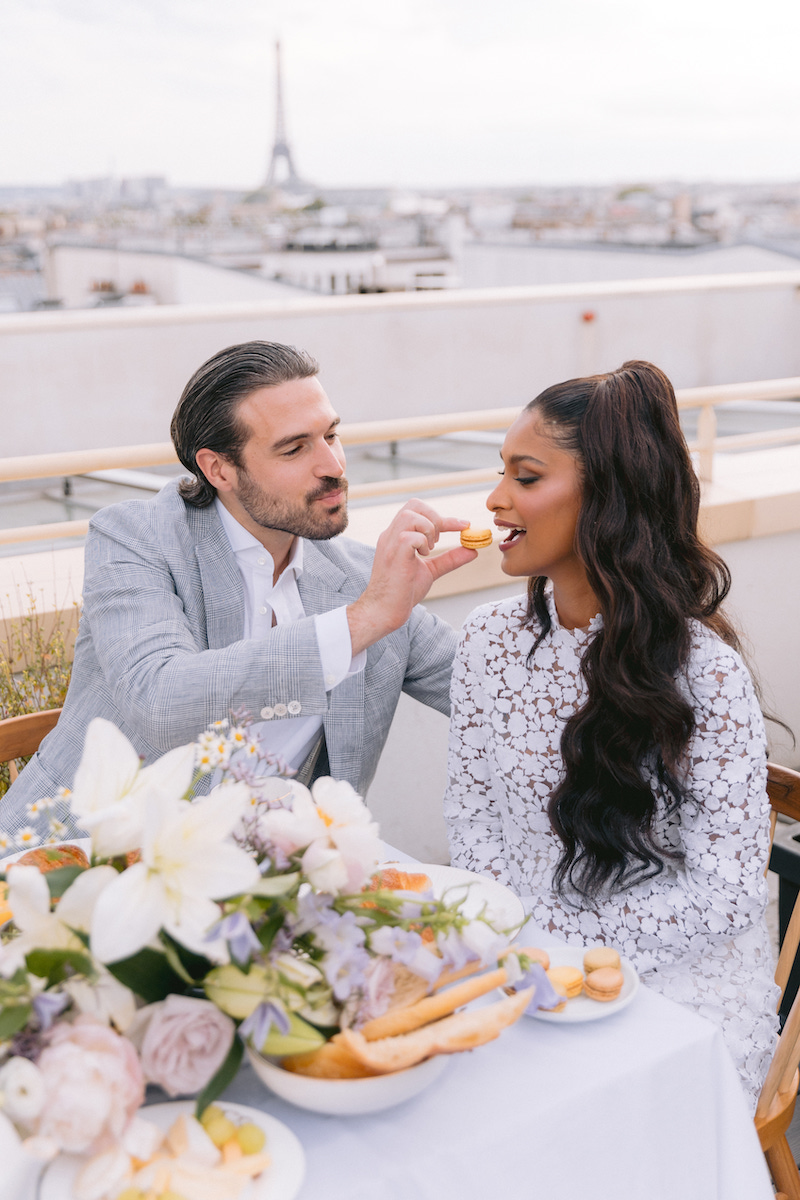 A man in a light suit feeds a woman in a white dress at an outdoor table with flowers and food, with the Eiffel Tower visible in the background.