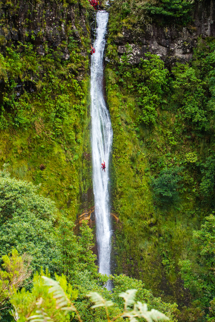 A person rappels down a tall, narrow waterfall in Madeira, Portugal, surrounded by lush green vegetation and steep rock walls.