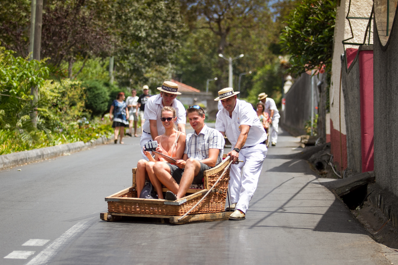 Two people sit in a wicker toboggan sled being pushed by two men in white uniforms and hats on a paved road in Madeira, Portugal, with more people in the background.