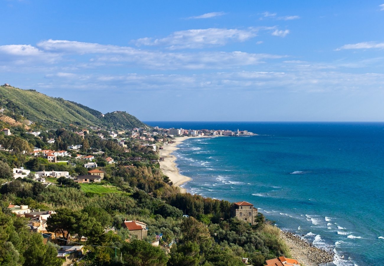 Coastal landscape with a seaside town, sandy beaches, green hills, and the blue ocean under a partly cloudy sky.