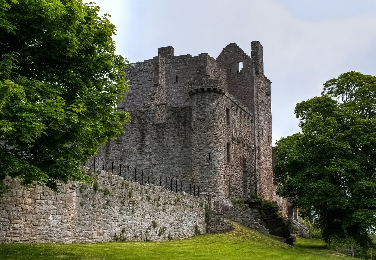 A stone medieval castle with crenellated walls, corner turrets, and a staircase, surrounded by green grass and trees.