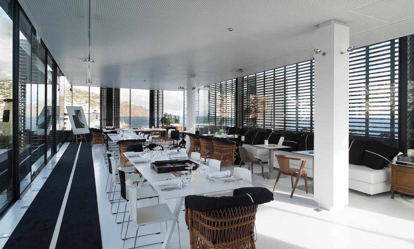 Modern restaurant interior with large windows, white floors and walls, black accents, contemporary furniture, and mountain views in the background. Tables are set for dining.