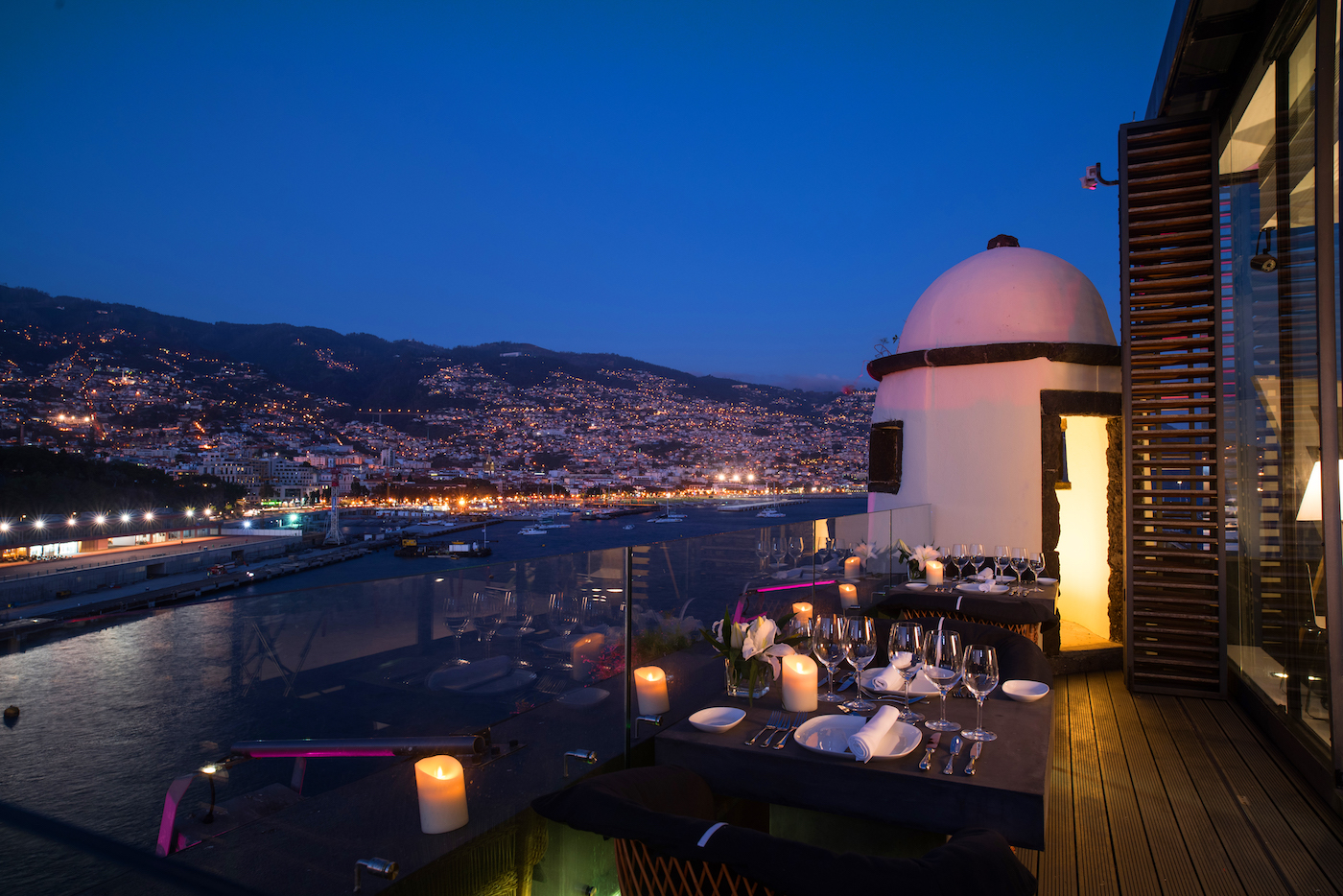 Rooftop restaurant at night overlooking a coastal city with mountains in the background, lit by city lights and candles on the tables.