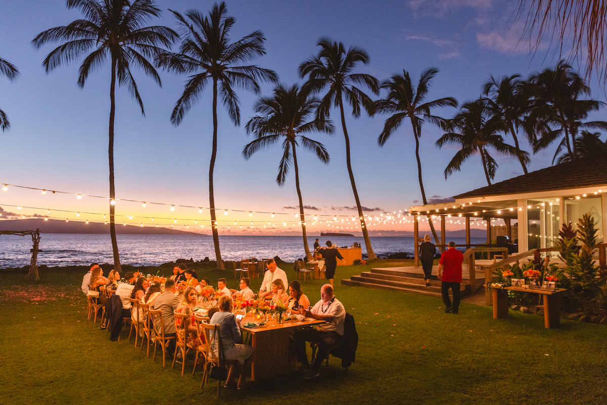 People dining outdoors at long tables under string lights near the ocean at sunset, with palm trees and a wooden structure in the background.